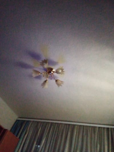 A ceiling light fixture with multiple frosted glass shades, shaped like bells or flowers, hangs from the ceiling. The fixture has a metallic gold-colored base. The ceiling appears to be textured and is lit, casting shadows from the light fixture onto it. To the right of the ceiling, a section of a wall or furniture with horizontal, striated lines in dark and light colors is visible, separated by a vertical white trim. A hint of a dark red, possibly wooden, surface is visible in the bottom right corner of the frame. The overall scene is indoors, likely a room, with ambient lighting. No people are visible. The image quality is somewhat grainy. No text is present. The location context of Synelnykove, Ukraine, does not provide specific visual cues within the image itself, such as landmarks or identifiable architectural styles.