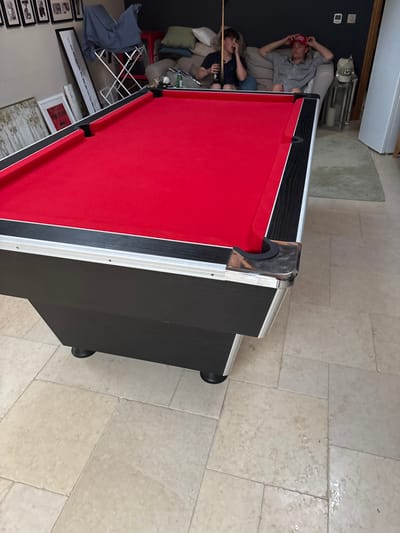 A red-felted pool table with black and silver trim dominates the scene. In the background, two people lounge on a couch, one in a red hat and the other holding a pool cue and what appears to be a drink. The table stands on a tile floor, with framed pictures lining a wall in the bottom left corner.