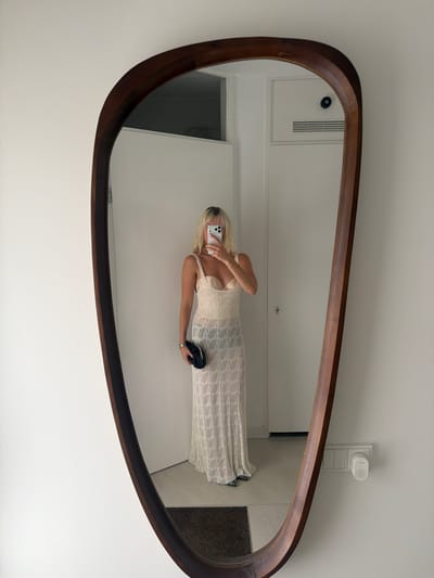 A woman with blonde hair is taking a mirror selfie. She is wearing a long, white, sleeveless dress with a textured pattern and a pearl necklace. She is holding a black clutch bag in her right hand and a smartphone in her left hand, with which she is taking the photo. The mirror is large and oval-shaped with a dark wooden frame. In the reflection, the woman is standing in a well-lit room with white walls. Behind her, a door and a ventilation grille are visible. To the right of the mirror, a wall-mounted electrical outlet with a small white device is visible. The overall scene suggests an indoor setting, possibly a dressing room or hallway. The image is captured from a low angle, as indicated by the orientation of the mirror and the reflection. There is no visible text in the image.