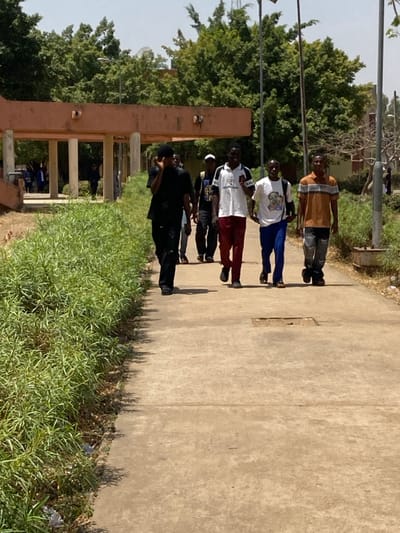 The image displays an outdoor scene in Minna, Nigeria, under bright daylight. A concrete pathway occupies the foreground and midground, exhibiting weathering and a central rectangular depression. Five adult males are walking along this path in a staggered line, generally moving towards the viewer.

From the left:
1.  An individual in black attire holds a dark object to their right ear, suggesting a phone call.
2.  A partially obscured individual in a dark shirt and light trousers follows.
3.  A male wearing a white polo shirt with "BALENCIAGA" branding, dark red trousers, and black footwear, carries a backpack and looks forward.
4.  A male in a white t-shirt featuring a graphic print, blue trousers, and dark footwear, also carries a backpack and looks forward.
5.  A male in a striped tan and white polo shirt, distressed grey trousers, and dark footwear walks on the right, looking forward.

Lush green shrubbery borders the left side of the pathway. In the mid-background, a terracotta-colored concrete structure with a covered walkway and supporting pillars is visible. Dense green trees dominate the background, with a white satellite dish partially visible on a distant building's roof above the trees. Additional indistinct figures are present further down the path and beneath the covered structure. Several vertical poles are scattered throughout the background.