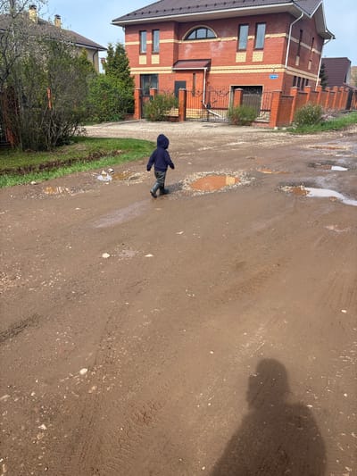 A young child, dressed in a dark hooded jacket and dark boots, is walking along a muddy street in Ферма, Russia. The street is riddled with puddles. A red brick house with a brown roof is on the left, fronted by a small lawn with sparse foliage and shrubbery. Tire tracks are visible on the muddy road surface.