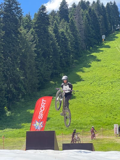 In Samokov, Bulgaria, a person wearing a helmet and protective gear is in mid-air on a mountain bike, performing a jump. The bike is tilted, with the front wheel lifted. The background consists of a lush green grassy slope dotted with trees, and a bright blue sky with some clouds. A red banner with the word "SPORT" and a colorful star logo stands to the left of the jumping cyclist. Further in the background, other cyclists are visible on the slope. The scene appears to be a downhill mountain biking event or training session.