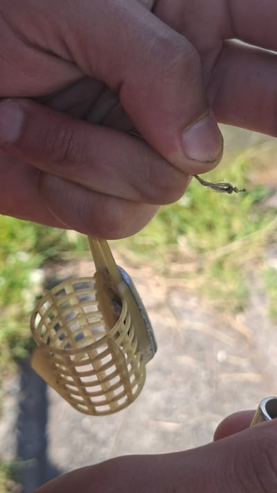 A close-up, outdoor shot shows a person's hands holding a fishing feeder. The feeder, made of light-colored plastic, has a basket-like structure with square openings. A metal component is attached to the feeder. The person's fingers, with visible dirt under the nails, are grasping the feeder and a thin, frayed cord. The background is out of focus, suggesting a natural environment with greenery and a paved surface. The image appears to be taken in or around Smalyavichy, Belarus.
