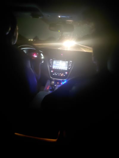 The image depicts the interior of an automobile at night, viewed from a rear passenger perspective in Radinovo, Bulgaria. A driver is partially visible on the left, positioned behind a steering wheel. A front passenger is also partially visible on the right. The vehicle's dashboard features a central, illuminated digital infotainment screen displaying a graphical user interface. Below it, the center console contains a gear selector and multiple backlit control buttons, illuminated with red and blue light. The windshield is visibly wet, presenting numerous water droplets or smudges, and reflects an extremely bright, centralized light source from outside the vehicle, likely oncoming headlights, which obscures the view forward. Less intense, distant lights are also visible through the upper section of the wet windshield. The overall scene is dark, with internal illumination primarily from the vehicle's controls and screen, complemented by a subtle amber light source visible in the lower right foreground of the cabin, possibly ambient lighting. The vehicle appears to be in motion.