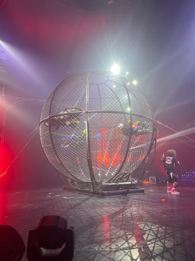 The image depicts a performance inside a large metal globe at a circus. Several motorcyclists are riding their bikes along the inner wall of the globe. A person with a distinctive afro hairstyle and a vest labeled "PHUKET CIRCUS" stands outside the globe, facing it. The stage is dimly lit with red and white spotlights casting dramatic shadows, including the geometric pattern of the globe's cage on the floor. The atmosphere suggests an ongoing, high-energy performance. The location is specified as Kathu, Thailand.