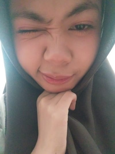 The media file is a close-up, angled selfie of a young woman wearing a dark hijab. Her head is tilted, and she is looking towards the camera with a playful expression, her right eye squinted and her left eye partially visible. Her lips are slightly parted, and she is touching her cheek with her left hand. The lighting suggests it might be indoors or during the day. The background is dominated by the dark fabric of the hijab, creating a simple and intimate setting. There is no visible text in the image. The overall mood is lighthearted and informal.