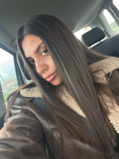A female individual is depicted in a close-up, front-facing view from inside a vehicle, presumably in the front passenger seat, within the city of Karnalovo, Bulgaria. She possesses long, straight, dark brown hair, brown eyes, and a neutral facial expression. The subject is attired in a brown, textured jacket featuring a cream-colored shearling-style collar and inner lining; a metallic zipper is visible on the garment's right side. The vehicle's interior components, including a gray headliner, which appears slightly sagged, and a gray rear seat headrest, are visible. Through the vehicle's left side window, residential buildings with red roofs, green vegetation, and an overcast sky are discernible. A yellow triangular sign with black text is partially visible through the rear window. The individual's gaze is directed towards the camera.