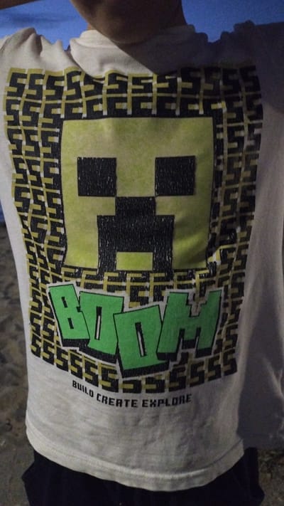 A close-up, slightly tilted shot shows a cream-colored t-shirt with a graphic design. The graphic features a pixelated green and black creeper face on the left, a large green word that appears to be "HIVE" on the right, and a repeating pattern of black and yellow shapes forming a border around the design. Below the "HIVE" text, in smaller vertical lettering, are the words "BUILD CREATE EXPLORE." The t-shirt is being worn, and the wearer's arm and shoulder are partially visible on the left side of the frame, along with a hint of a blue garment underneath. The t-shirt is laid out on a dark surface, possibly a floor or ground, with a dark blue or black garment visible beneath the right side of the t-shirt. The lighting is dim, suggesting it is nighttime or indoors. The overall impression is of a casual, possibly gaming-themed t-shirt. There are no explicit clues about the time of day, weather, or specific location within Perea, Greece, beyond the provided context. The image does not convey any discernible emotions.