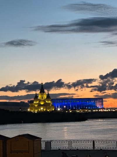 The image depicts a twilight scene in Nizhny Novgorod, Russia, showcasing the confluence of the Oka and Volga rivers. In the foreground, a tranquil body of water reflects the dim light, with a metal railing and the tops of small wooden structures visible along the embankment. Across the water, a prominent church, illuminated with warm yellow lights, stands out against the darkening sky. To its right, a large stadium is bathed in vibrant blue lights, creating a stark contrast. The sky transitions from a soft, pale blue at the top to hues of orange and pink near the horizon, interspersed with dramatic dark clouds. The overall atmosphere suggests a serene evening in the city, with architectural landmarks illuminated for evening viewing.