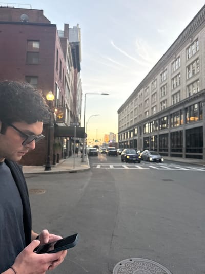 The image depicts an urban street scene in the downtown area of Boston, Massachusetts, United States, during what appears to be late afternoon or early evening.
In the foreground, slightly left of center, a male individual with dark, curly hair and dark sunglasses is seen in profile, looking down at a dark-colored smartphone held in both hands. He wears a dark t-shirt and a dark open jacket or vest. A light-colored bracelet is visible on his left wrist. Part of a circular metal manhole cover with a geometric pattern is visible on the asphalt pavement in the lower right.
The street, paved with asphalt, extends into the background, showing white crosswalk markings in the mid-ground. Multiple cars are present on the street, traveling away from the viewer; some have illuminated headlights. The street is flanked by multi-story buildings. On the left, a red-brick building with visible windows features a prominent street lamp with a glowing yellow light and a "ONE WAY" sign below it. Some decorative, warm-toned lighting is visible near an entrance on this building. On the right, a lighter-colored, possibly limestone or light brick, multi-story building with numerous large windows on its lower floors and smaller windows on upper floors lines the street, reflecting the sky and interior lights.
The sky in the background is light blue with a warm, yellowish glow near the horizon, and faint white contrails are visible. Distant urban architecture and an illuminated sign are discernible further down the street.