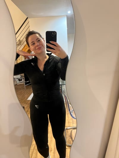 A woman in a black athletic outfit poses in front of a uniquely shaped mirror, holding her phone up to take a photo of herself. She is smiling slightly. The reflection shows a room with a chair in the background, along with wooden flooring and walls with white accents. The wavy edge of the mirror slightly distorts the view.