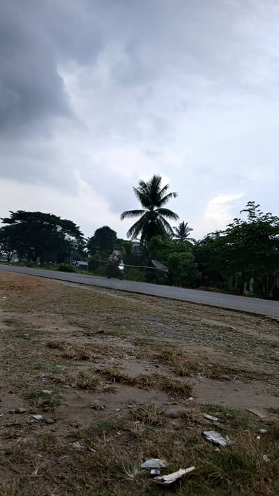 The image depicts a rural scene in Sumbokrayek, Indonesia, under a cloudy sky. A paved road runs horizontally across the mid-ground, bordered by sparse dry grass and gravel on the left and lush greenery on the right. A motorcycle, with its headlight illuminated, is in motion on the road, appearing blurred due to movement. Behind the road, a dense line of trees and vegetation is visible, including a prominent coconut palm tree. In the background, more trees and some houses are discernible. The foreground consists of a dry, patchy grassy area with scattered rocks and some litter. The overall atmosphere suggests an overcast day in a tropical or subtropical environment.