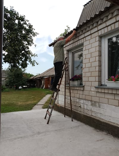 An adult male wearing a striped shirt and a cap is standing on a ladder, reaching towards the roof of a light-colored brick building. The building has windows with flower boxes. In the background, there is a grassy yard with trees and other structures, suggesting a rural or suburban setting. The image appears to be taken in Hamarnia, Ukraine. The overall scene depicts a person engaged in outdoor maintenance.