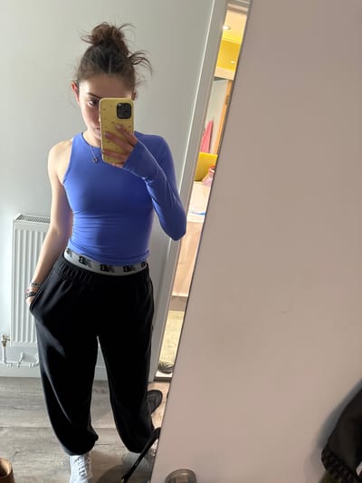 A young woman is standing in front of a mirror, taking a selfie with a yellow smartphone. She is wearing a blue long-sleeved top and black sweatpants with a New Balance logo waistband. Her hair is pulled back in a messy bun. The reflection in the mirror reveals a room with a radiator, wooden flooring, and other miscellaneous items out of focus in the background. The location of this image is Hull, United Kingdom.