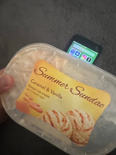 In Shotton, United Kingdom, a close-up shot captures a tub of "Summer Sundae" ice cream with "Caramel & Vanilla flavour ice cream with Caramel ripple" branding. The lid is off, and the ice cream, with visible caramel swirls and two scoops, is partially shown. A smartphone is propped up inside the empty side of the tub, its screen displaying the "App Library" with icons for WhatsApp, Instagram, and other apps. The background is a dark, textured surface, possibly a sofa or carpet. The image is taken from a slightly high angle, focusing on the ice cream and the phone.