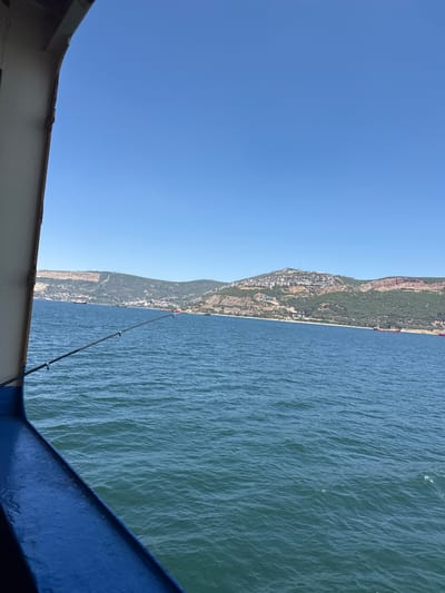 An eye-level shot captures the view from the side of a vessel on a clear, sunny day. The deep blue sky dominates the upper half of the frame, meeting a distant, green and tan-colored hill that slopes down to the water. A small town with white buildings is visible clustered on the side of the hill. Several red cargo ships are anchored in the blue water. In the foreground, a fishing rod extends from the left side of the boat, angled towards the distant shore. The boat's blue railing is prominent in the lower left corner, with a white section of the boat's hull visible above it. The water has small, gentle ripples.