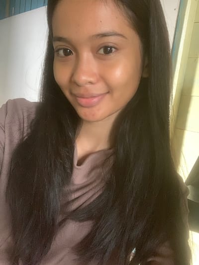 The image is a close-up, waist-up selfie of a young woman with long, dark hair. She is wearing a mauve, cowl-neck top. Her skin appears smooth with a natural glow. Her eyes are dark and she is looking slightly off-camera, with a faint smile. The background is out of focus, but appears to be an indoor setting with a white wall to the left and yellowed wooden paneling to the right, suggesting an interior space possibly in Sungai Meriam, Indonesia. The lighting creates some shadows and highlights on her face and hair. There are no other people or significant objects visible in the frame.