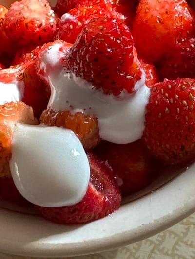 The image is a close-up, overhead shot of a bowl filled with fresh strawberries and dollops of white cream. The strawberries are ripe, vibrant red, with visible seeds. The cream appears thick and smooth, partially obscuring some of the fruit. The bowl itself is a light beige or off-white color, with a slightly textured surface. The background is out of focus, but suggests a patterned surface, possibly a tablecloth or placemat. No people, actions, or interactions are depicted. The image is a food still life. There is no information in the image to suggest a location in Biysk, Russia.