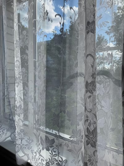 The image shows a close-up of white lace curtains with a floral pattern, hanging in front of a window. Through the translucent fabric, the blurred shapes of trees and a blue sky with white clouds are visible. The lighting suggests it is daytime, with sunlight filtering through the curtains. The context of Naro-Fominsk, Russia, is not directly evident from the visual elements of the image itself.