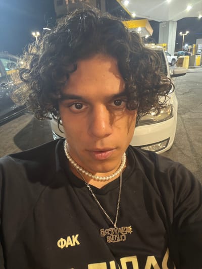 The media file shows a close-up, slightly tilted, selfie of a young person with curly dark hair. They are wearing a black sweatshirt with a pearl necklace and a silver chain with a pendant. The sweatshirt has some visible text on it, including "ΦΑΚ" on the right side and a more elaborate, possibly jeweled, pendant with text that is difficult to decipher.
The background appears to be a brightly lit outdoor setting at night, possibly a gas station or a similar commercial area. There are bright lights and structures visible, including what looks like a white vehicle in the background. The overall lighting suggests it is nighttime. The location context provided is Netanya, Israel. The subject's expression is neutral, and the image is likely a casual personal photograph.