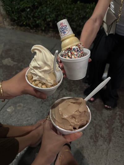 Here's a detailed description of the image:
The image depicts three people holding ice cream.
1. The person on the left is holding an ice cream cone in a cup that says "Joy of the United States of America 250". The cone is covered in sprinkles.
2. The person at the bottom is holding a cup of ice cream with whipped cream and nuts on top. A white plastic spoon is present.
3. The person on the right is holding a cup of chocolate ice cream. A white plastic spoon is present.
The photo is taken at night. In the background, there's a sidewalk. The people are standing in a casual setting. The photo is most likely taken in the city of Boca Raton.