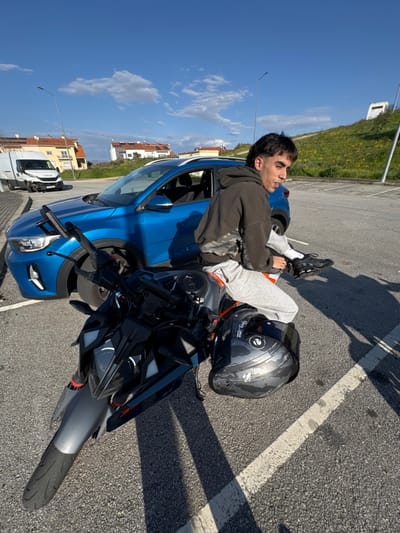 The image depicts an outdoor scene in Mirandela, Portugal, set within a paved parking area under a clear blue sky with scattered cumulus clouds. In the foreground, a male individual with dark hair styled in a mullet sits on a grey, black, and orange sport-naked motorcycle. He is wearing a dark hooded sweatshirt, light grey sweatpants, and black athletic shoes with white socks, looking slightly to his left. A scuffed black full-face motorcycle helmet rests on the motorcycle's tank or seat area. Behind the motorcycle, a blue compact crossover SUV, consistent with a Kia Stonic model, is parked. Further in the background, a white Iveco commercial van is visible on an adjacent road, its front passenger-side bumper appearing damaged or dislodged. Residential buildings with light-colored facades, red-tiled roofs, and visible solar panels are in the mid-ground, while a grassy, unkempt slope rises on the right side of the background. Streetlight poles are present against the sky.