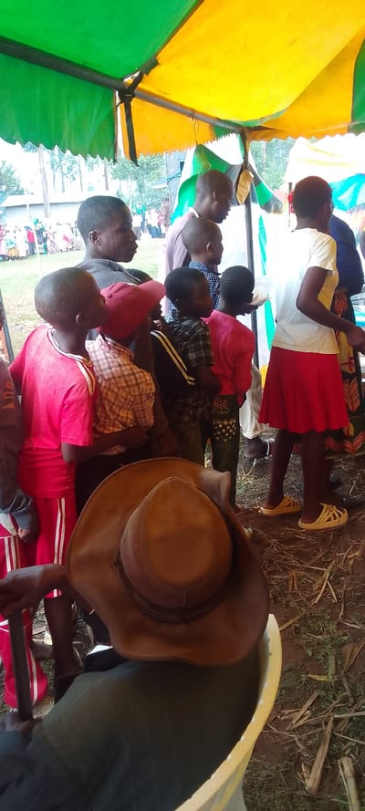 The image shows a group of people gathered under a yellow and green striped tent. The people are mostly children, all wearing casual clothes, with some adults. One of the children is wearing a red shirt with white stripes and red shorts with white stripes. Another child is wearing a plaid shirt and brown pants. There is also a woman wearing a red skirt and a white top. The tent is located in a grassy area with dried grass and leaves on the ground. The ground is brown and appears to be dirt. The people are standing in a line. One adult is wearing a brown cowboy hat. The image is taken from a low angle. The coordinates provided do not correspond to a location in Kenya. Kenya is located in East Africa. Lubao is a municipality in the Philippines.