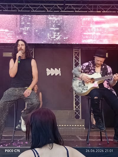 The image shows a live music performance on an outdoor stage. Two men are the primary subjects. The man on the left is seated and singing into a microphone, wearing a black tank top and patterned pants. He has long dark hair and a mustache. The man on the right is also seated, playing an acoustic guitar. He wears a black fedora, a patterned shirt with pink and black elements, and black pants. His guitar is decorated with drawings and text. In the background, a large screen displays a pink and white shimmering light pattern. Another person is partially visible seated behind the singer. The stage has metal scaffolding and lights. The overall atmosphere appears to be that of a daytime outdoor concert. Visible text includes "POCO C75" and "26.06.2026 21:01" in the lower corners of the image. The location context provided is Wrocław, Poland, though this is not directly evident from the image itself.