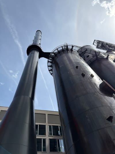 The image shows a low-angle view of industrial structures in Esch-sur-Alzette, Luxembourg. A towering, dark gray or black cylindrical structure dominates the frame, composed of stacked tanks or sections. Metal grates, ladders, and platforms connect the different levels. Adjacent to the industrial structure, a portion of a modern building with a grid of windows is visible. The sky is light blue with faint contrails and a faint halo effect around the sun.