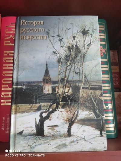 The image shows a close-up of several books stacked on a shelf. The main focus is on a book with a painting of a winter scene on its cover. The painting depicts bare trees with nests in their branches, a snow-covered landscape, and a church spire in the background. The title of this book, "История русского искусства" (History of Russian Art), is printed vertically in gold lettering on the left side of the cover.
Above this book, a green book with gold and red decorative elements is visible, with some Cyrillic text. Below the main book, another red book with gold lettering is partially visible, displaying Cyrillic text that appears to be a title or author's name. The overall setting appears to be a bookshelf, likely in a home or library, given the presence of multiple books. The lighting suggests an indoor environment, and the scene is still, with no people or overt activity taking place. The presence of the text and the book titles suggests a cultural or educational context. The watermark "POCO X3 PRO | ZDANNATS" is also visible on the right side of the image.