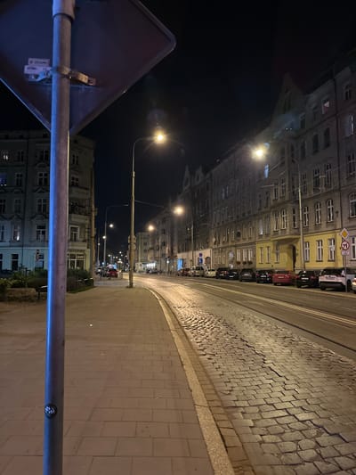 A nighttime street scene in Wrocław, Poland. The image is oriented vertically, showing a perspective looking down a cobblestone street lined with ornate, multi-story buildings. Cars are parked along the side of the street, and streetlights illuminate the scene.
The buildings on the left are detailed with many windows and classical architectural elements. Some windows are lit, suggesting occupancy. The street itself is paved with cobblestones, which appear worn and uneven in places. To the right of the cobblestone section, there is a sidewalk made of rectangular paving stones.
In the foreground, a metal pole and the back of a diamond-shaped sign are visible. The sign is dark and triangular, with a metal bracket attaching it to the pole. The pole is also illuminated with a blueish light, possibly from another light source not directly in view.
The time of day is clearly night, as indicated by the dark sky and artificial lighting. The weather appears to be clear, with no visible precipitation. The scene evokes a sense of urban ambiance at night, with the architecture suggesting an older European city. No people are prominently visible.
Visible text includes a diamond-shaped traffic sign with a red circle and diagonal line, indicating a prohibition. There is also some text on a sticker on the metal pole, which is blurry but appears to say "ZOOM" and "CE". On the building on the right, partially obscured by the foreground pole, there appears to be a sign with Cyrillic or similar script, possibly a shop name.