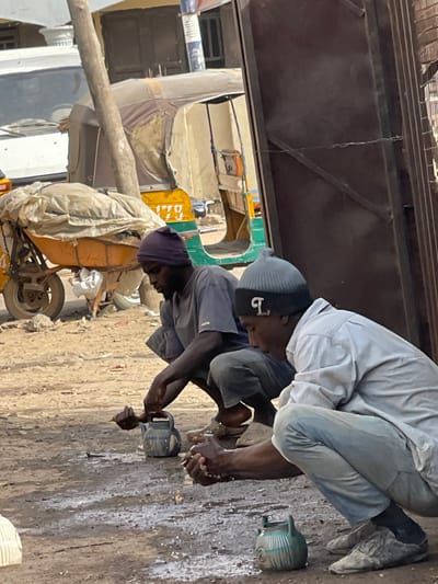This image captures an outdoor scene in Zaria, Nigeria, featuring two adult males engaged in handwashing on an unpaved, dusty ground surface. White foamy residue and water splashes are visible on the ground directly in front of both individuals.
The man positioned on the left, with dark skin, is wearing a dark purple knit beanie, a short-sleeved grey t-shirt, and dark trousers. He is squatting, utilizing his left hand to pour water from a blue, patterned metal kettle onto his right hand, which is held cupped over the ground.
The man on the right, also with dark skin, wears a grey ribbed knit beanie with a black band featuring a white 'L' emblem, a light blue long-sleeved button-up shirt, and light-colored trousers. He is also squatting, holding his cupped hands over the ground, appearing to rinse them. A light green, patterned metal kettle rests on the ground in front of him.
In the background, a yellow and green three-wheeled auto-rickshaw (Keke Napep) with the number "1770" visible on its side is parked. A white vehicle is partially visible further to the left. A yellow wheelbarrow covered with a white tarp is situated behind the left man. A vertical wooden pole stands in the mid-ground. On the right side of the frame, a dark brown metal gate or door, secured with a wire latch, is visible. Buildings are faintly discernible in the far background. The scene is illuminated by bright daylight.