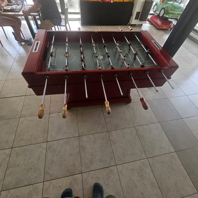 The image depicts a foosball table in an indoor setting, likely a public space given the tiling and visible background elements. The foosball table is predominantly red with a green playing surface and metallic rods with player figurines. There are at least two people partially visible: one person is seated at a table to the left, and another person is standing, their feet in dark blue crocs are visible at the bottom of the frame. The background reveals a tiled floor, a large black structure, and a green kiddie ride shaped like a car. The scene appears to be well-lit, suggesting daylight or artificial lighting. The overall impression is that of a casual recreational area within a larger establishment.