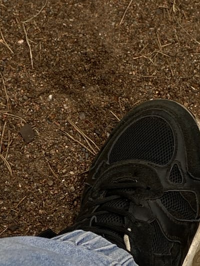 An overhead shot shows a black sneaker with white laces on a person's foot, positioned over a ground surface composed of small pebbles and dirt. The sneaker is a dark color, possibly black or dark gray, with visible mesh sections on the toe and sides. The laces are black, with a small white accent on one of them. The person is wearing light blue jeans, visible at the bottom left of the frame. The ground is a mixture of brown dirt and small, multicolored pebbles, with a few dry twigs or grass stalks scattered across it. The lighting suggests an outdoor environment, possibly during the day. The image does not provide any specific landmarks or context to determine its location within the city of Baltiiskoe, Russia.