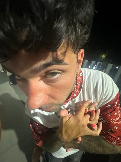 A close-up, high-angle, dimly lit outdoor shot shows a young person with dark, messy hair, brown eyes, and light skin. They are wearing a white t-shirt with a red bandana-like pattern on the sleeves and collar. The person's hands are clasped together, and their fingernails are painted bright pink. A silver chain with a pendant is visible around their neck. The background features dark shadows, a hint of a light-colored railing, and some blurred lights in the distance, suggesting it is nighttime. The ground appears to be paved. The image does not provide enough information to determine the specific location in El Masnou, Spain.