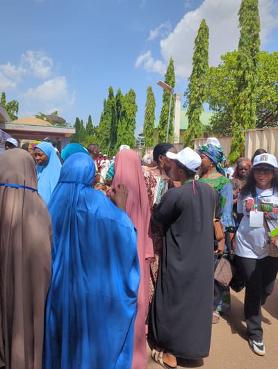 In Ungwan Liman, Nigeria, a group of people are gathered outdoors, likely at an event or gathering. The group appears to be predominantly women, some wearing head coverings like hijabs and others wearing white caps. Garments include various shades of blue, pink, black and brown. Some people are wearing white t-shirts and black trousers. The background includes trees, buildings, and a blue sky with clouds.