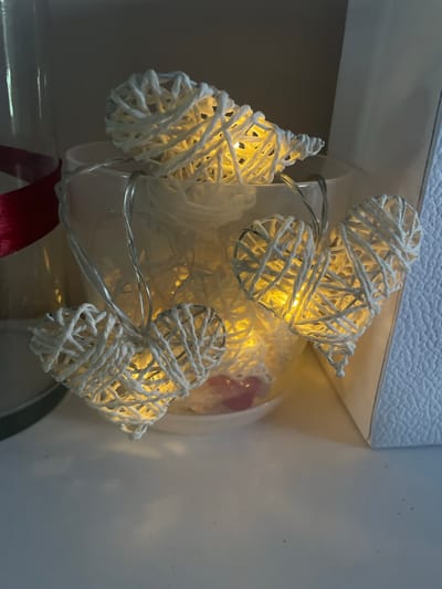 A string of heart-shaped decorative lights made of white woven material is displayed inside a clear glass container, possibly on a shelf in a residential setting in Brovary, Ukraine. The lights are illuminated with a warm yellow glow, casting a soft light on the surrounding objects. To the left of the container, a red ribbon is partially visible, suggesting a gift or decorative element. The background is dimly lit and out of focus, with a textured white surface on the right side of the image. There are no people or animals present in the scene.