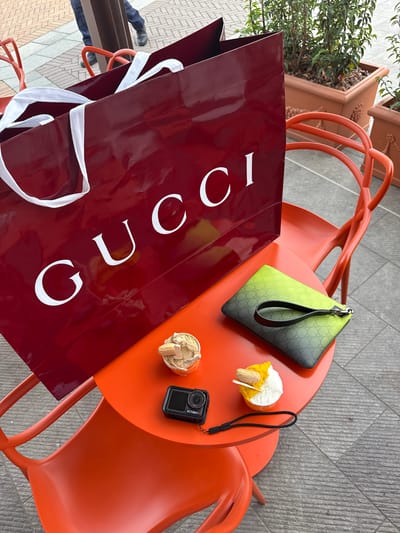 A high-angle shot shows an orange outdoor table with an adjacent plant container and gray-tiled ground. The table holds a red Gucci shopping bag, a green and black Gucci pouch, two small cups of dessert or ice cream, and a small black action camera (Action 5). The camera is in the lower-right quadrant. The items are arranged as if recently placed on the table. A person's legs are partially visible in the lower-left corner. This scene is located in Noventa di Piave, Italy.