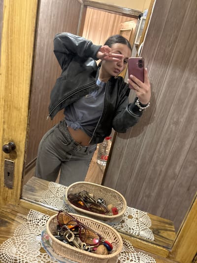 A woman is taking a selfie in a mirror indoors. She is wearing a gray t-shirt tied at the waist, gray jeans, and a black leather jacket. She is making a peace sign with her left hand. A bracelet is visible on her left wrist. She is holding a pink smartphone in her right hand. Behind her, there is a wood-paneled wall. In the mirror's reflection, a partially visible water bottle is on the floor. To the right, on a wooden surface adorned with lace, are two wicker baskets filled with items like keys and sunglasses. The setting is implied to be in Radomir, Bulgaria, although there are no visual cues in the image to confirm the location.