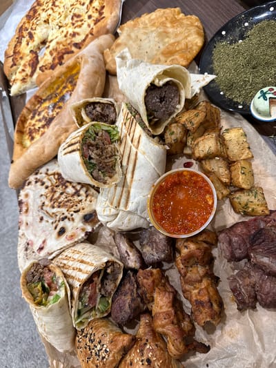 This is an overhead shot of a platter with various Middle Eastern foods. The platter includes wraps containing meat and vegetables, a variety of bread including naan and pita, fried potatoes, cooked meat, and a container of red sauce. A small bowl filled with herbs sits on the upper left of the platter. The food is presented on a sheet of brown paper, and the platter is on a dark, wooden surface. There is no indication of the city of Philadelphia or any human presence in the image.