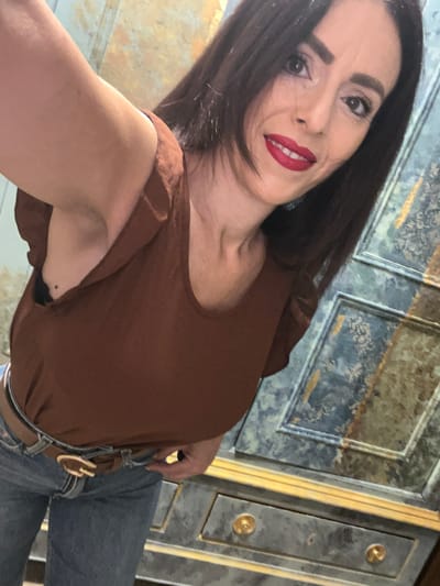 A woman with dark hair and red lipstick is facing the camera. She's wearing a brown top and jeans with a brown belt. The background features a decorative cabinet or wall panel, possibly in a historical or ornate style. The location in the city of Konz, Germany, is unknown from the image.