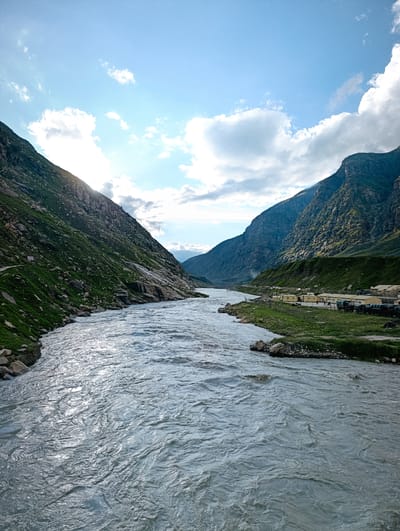 The image depicts a wide, fast-flowing river cutting through a valley in Koksar, India. Steep, rocky mountains covered in sparse green vegetation flank the river on both sides. The sky is bright blue with scattered white clouds, and the sun is partially obscured by the mountains on the left, casting a light glare. On the right bank of the river, there is a settlement with several buildings that appear to be made of beige-colored structures, possibly temporary shelters or barracks, with a row of dark-colored vehicles parked in front. The river's surface is turbulent, with visible ripples and reflections of the sky. A narrow, dirt path is visible on the grassy slope of the left mountain. The overall scene conveys a sense of a remote, natural landscape with a small human presence.