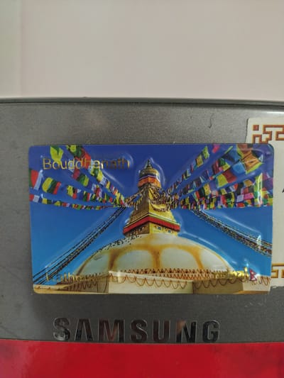 A close-up shot shows a refrigerator with a magnet depicting the Boudhanath stupa, located in Kathmandu, Nepal, attached to it. The magnet is colorful and features the iconic Buddhist stupa, prayer flags, and a blue sky backdrop. The "SAMSUNG" brand name is visible on the refrigerator door. The rest of the refrigerator is gray and red in color.