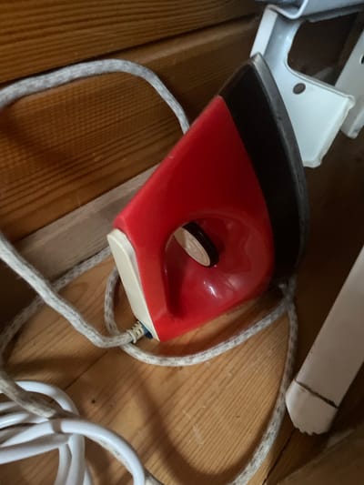 The image shows a close-up of a red and black electric iron with a white cord lying on a wooden surface. The iron is partially obscuring a white metal object. The wooden surface appears to be part of a shelf or floor. The background is dark and out of focus. There are no people or animals in the image. The overall scene suggests a storage area. The image does not provide any information that would allow for its location to be specified as being in the city of Турово, Russia.