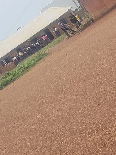 This image depicts an outdoor scene in front of a building, likely a school or community center, in Gumel, Nigeria. A group of young people, some appearing to be students, are gathered outside. One individual, dressed in a black t-shirt with the number "99" and grey shorts, stands near a bicycle. Another person in a yellow t-shirt is also visible. The ground is a reddish-brown dirt path. In the background, the building has a light-colored, possibly corrugated metal roof and brick walls. The sky is overcast. The overall impression is of a casual gathering or a moment in the daily life of the community.