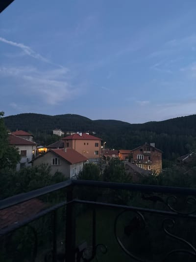 The image depicts a dusk view of the city of Pernik, Bulgaria. The scene is framed by a dark, ornate metal railing in the foreground, suggesting a balcony or patio. Beyond the railing, a residential area is visible with several multi-story buildings with red-tiled roofs and light-colored facades. One building, constructed of red brick, stands out with visible windows and a chimney. In the background, a dense, dark green forest covers rolling hills, characteristic of the natural landscape surrounding Pernik. The sky is a gradient of deep blue, transitioning to lighter shades near the horizon, with a few wispy clouds and a faint contrail visible. The ambient light suggests it is either early evening or late dusk, with some artificial lights illuminated within the buildings. No people are visible in the image.