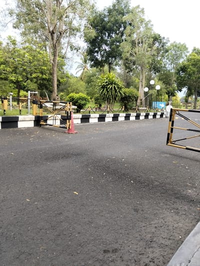 The image shows a barrier gate at the entrance to a property in Tambontunong, Indonesia. The gate is made of yellow and black bars and is partially open. A red traffic cone is placed in front of the gate. The road is asphalt and has some fallen leaves on it. In the background, there are trees, bushes, and lampposts. The sky is overcast. The scene appears to be a quiet and serene environment.