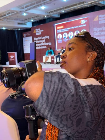 An individual, identified as a female, is positioned in the foreground, facing right in profile, operating a professional video camera. Her left arm supports the camera system, which includes a black mirrorless camera with a lens, mounted on a black handheld gimbal stabilizer. A rectangular, dark blue accessory is attached to the camera's hot shoe. She wears a dark grey shirt with a lighter grey or white shoulder stripe and an intricate dark patterned design. Her hair is styled in orange and black braids, with dark sunglasses resting on top of her head. A rose-shaped earring is visible on her left ear.

The background reveals a large indoor event space in Itirin, Nigeria, specifically a conference or forum setup. A stage is prominent, featuring a large red backdrop with white text and logos. Visible text includes "Nigeria Solar Forum" and "Nigeria's Leading Gathering of Renewable Energy Stakeholders." A screen on the left of the stage displays "The Solar Road Map To Nigeria's Energy Independence" alongside multiple profile images of individuals. Two individuals are seated on the stage: one female in a teal dress and one male in a suit. Overhead lighting fixtures are visible on the white ceiling. Partial views of other attendees are visible in the blurred foreground, suggesting an audience setting.