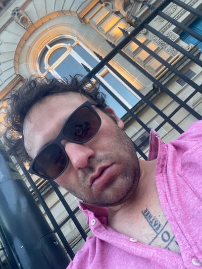 The image shows a close-up, high-angle selfie of a man wearing sunglasses and a pink collared shirt. He has dark, curly hair and stubble. A tattoo is visible on his chest, with the word "FATHER" written vertically. In the background, there is a building with a stone facade and a large arched window. A black metal fence is in the foreground, obscuring part of the building and the man's body. The lighting suggests it is either early morning or late afternoon, with sunlight casting shadows on the building. The location is identified as Armagh, United Kingdom.