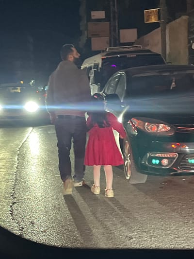 The image displays an urban street scene at night in Al-Kiswa, Syria. An adult male and a young female child are walking away from the camera, positioned on the right side of a wet, reflective asphalt road.

The adult male, located to the left of the child, wears a dark jacket, dark trousers, and light-colored shoes. A black face mask is visible pulled down around his chin. He has dark hair and a beard. His right arm is extended, and his hand appears to be holding the left hand of the child.

The young female child, situated to the right of the adult male, wears a red dress, white tights, and light-colored strapped shoes. She has long, dark hair.

To their right, a dark-colored sedan is parked with its driver's side door open, extending towards the street. The sedan's headlights are illuminated, featuring distinct green-tinted elements. Behind the sedan, a white van or minibus with a visible roof rack is partially obscured. In the far left background, bright headlights from an approaching vehicle cast a significant glare. Further background details include indistinct buildings or walls, a utility pole, and several rectangular objects affixed to a structure. The asphalt road surface shows visible cracks and reflects ambient light, suggesting it is wet.